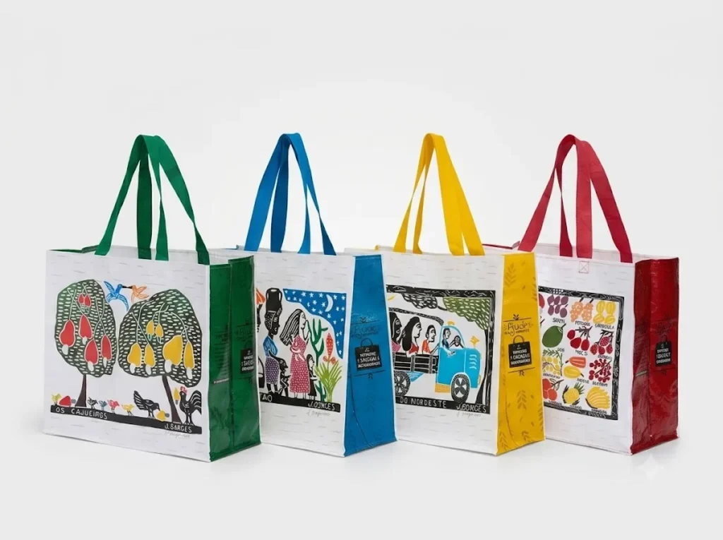 Quatro sacolas de ráfia brancas alinhadas exibindo as obras de arte em xilogravura de J. Borges com acabamentos laterais nas cores verde, azul, amarela e vermelha.