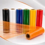 Bobinas de filme stretch colorido nas cores preto, branco, azul, verde, amarelo, vermelho e laranja com filme laranja estirado em fundo de estúdio.